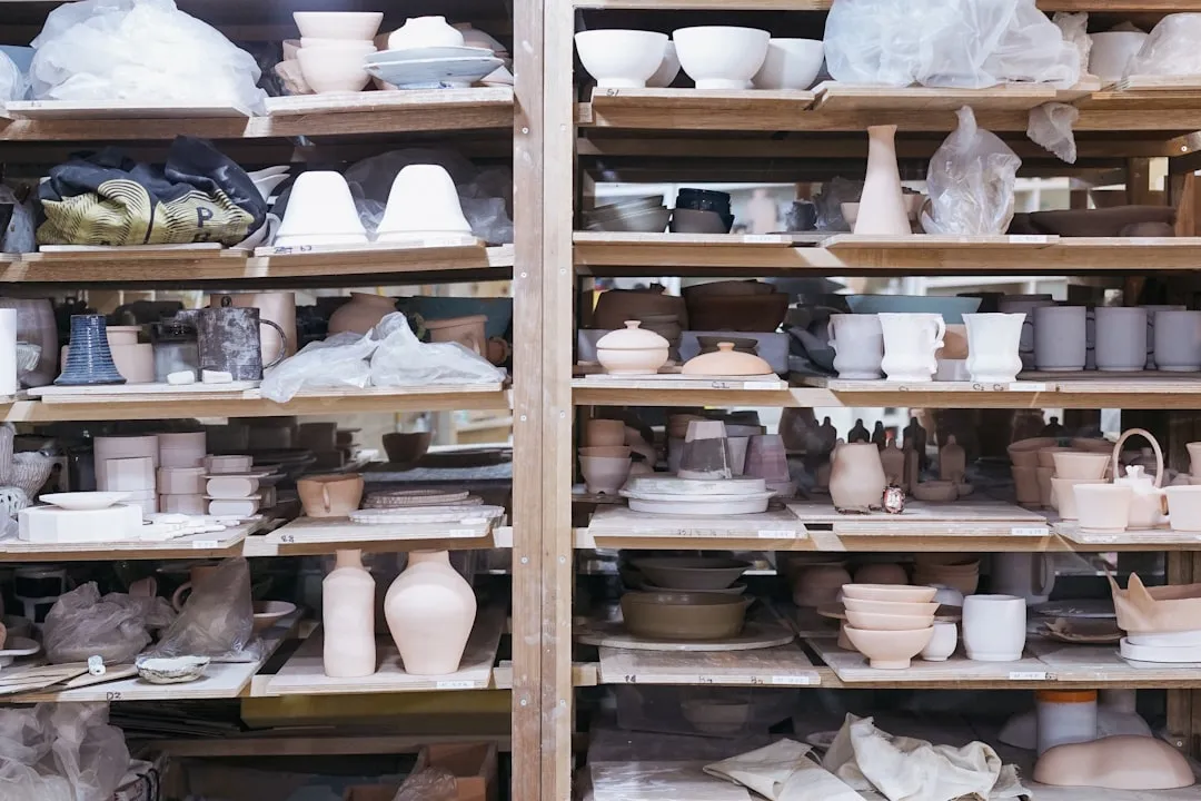 Pièces de vaisselle en céramique non émaillée stockée sur des étagères en bois, préparée pour la cuisson ou la décoration dans une fabrique de poterie artisanale.